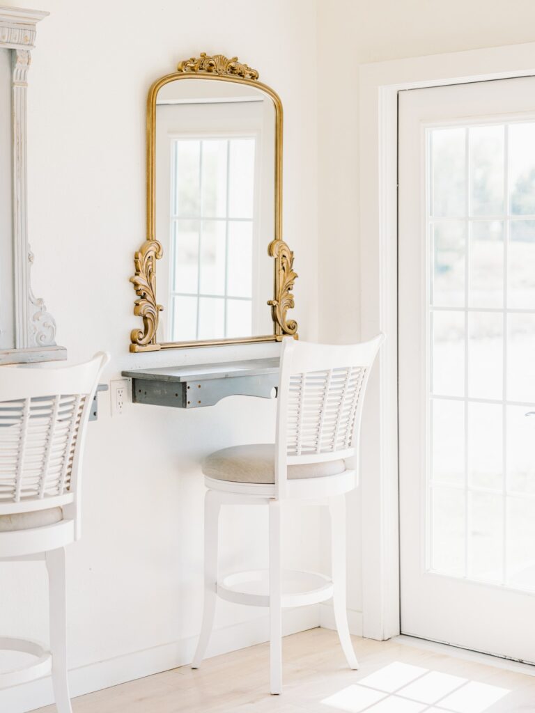 Makeup station at The Grand Ivory bridal cottage with white chair in front of mirror with gold trim, next to full length window.