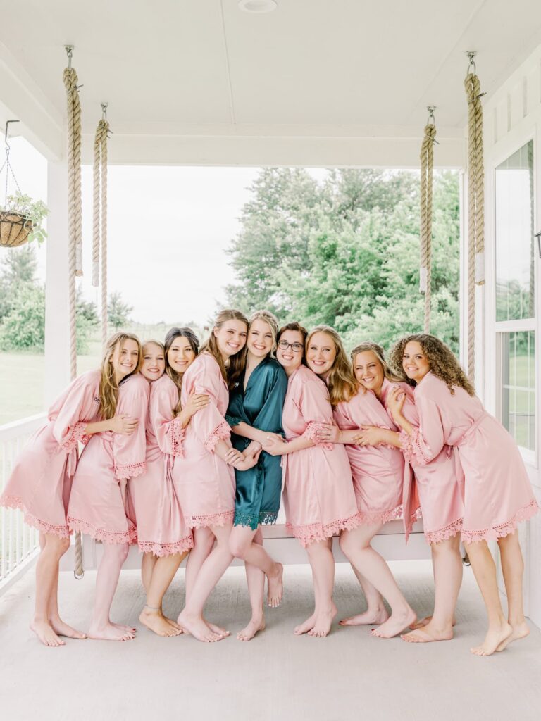 Bride and bridesmaids in satin robes hugging on porch in front of swing.