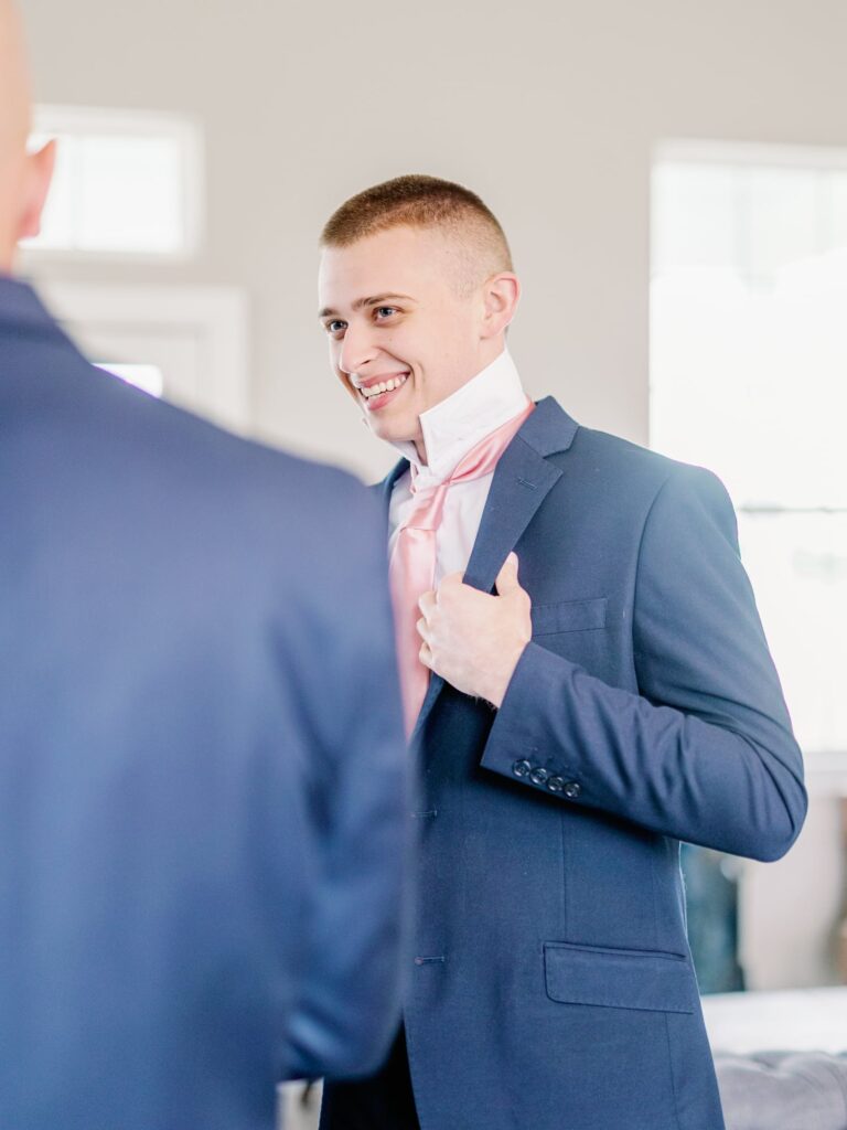 Groom getting dressed in a navy suit with blush tie smiling at groomsmen.