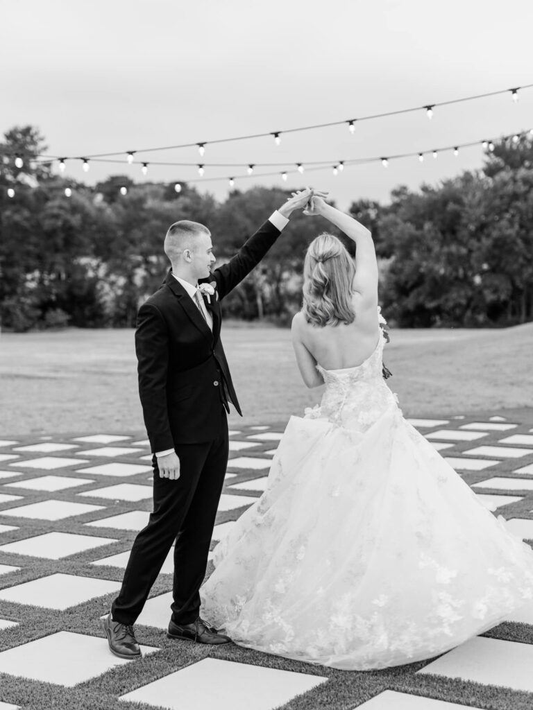 Couple twirling underneath string lights and grass lattice in wedding attire.