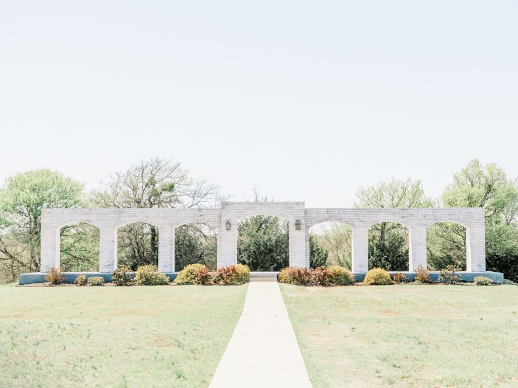 Ceremony space with white arches and greenery in front, trees in the background for weddings.