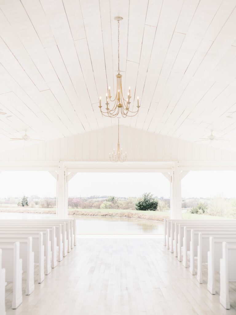 Open air chapel with two gold chandeliers and white church pews at wedding venue.