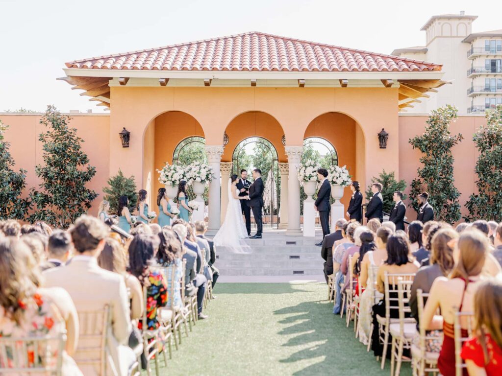 Bride and groom holding hands during wedding ceremony surrounded by friends and family at the Rosewood Mansion venue in Dallas.