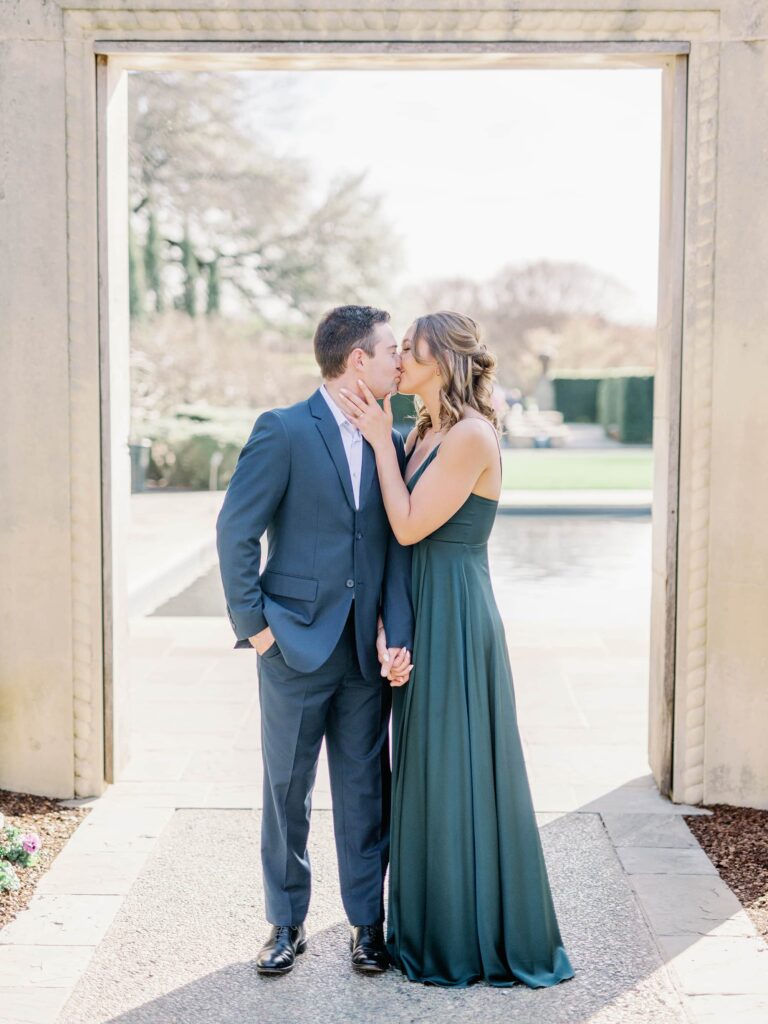 Groom in blue suit and bride in emerald floor length gown together celebrating their engagement by taking photos at the Dallas Arboretum.