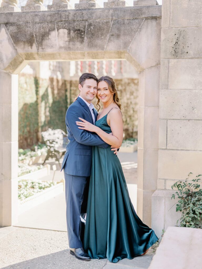 Engaged, brunette couple in formal attire hugging and smiling while at the Dallas Arboretum.